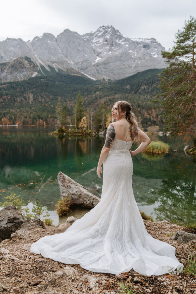 A woman in a white wedding dress poses during her Germany elopement in front of an emerald green lake Eibsee, which is surrounded by fall colors and snow capped mountains