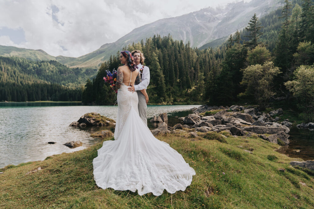 A bride in white wedding dress holds her husband on top of a grassy green hill surrounded by lush green mountains and a turquoise lake as they elope in Germany