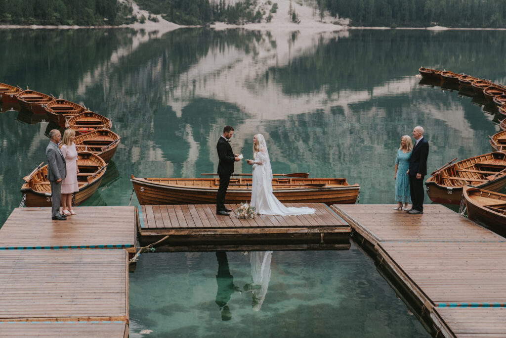 A couple stands on a wooden dock with parents on each side, reading their vows on their wedding day during a small minimony celebration in the Dolomites