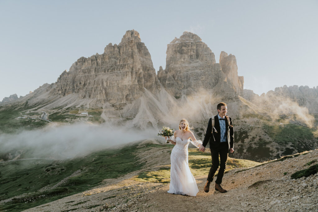 A couple wearing wedding attire walks holding hands and laughing during their elopement day with the dramatic Dolomites mountains in the background