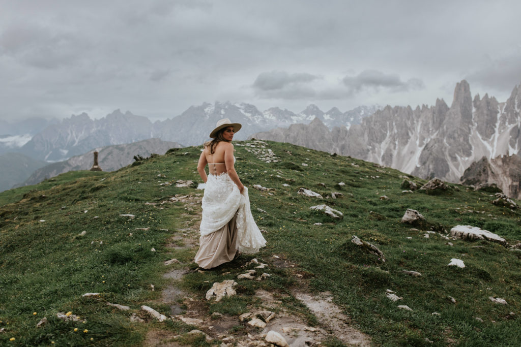 a bride walks in her wedding dress along the Tre Cime hiking trail on her elopement day, with the Cadini di Misurina peaks far in the background