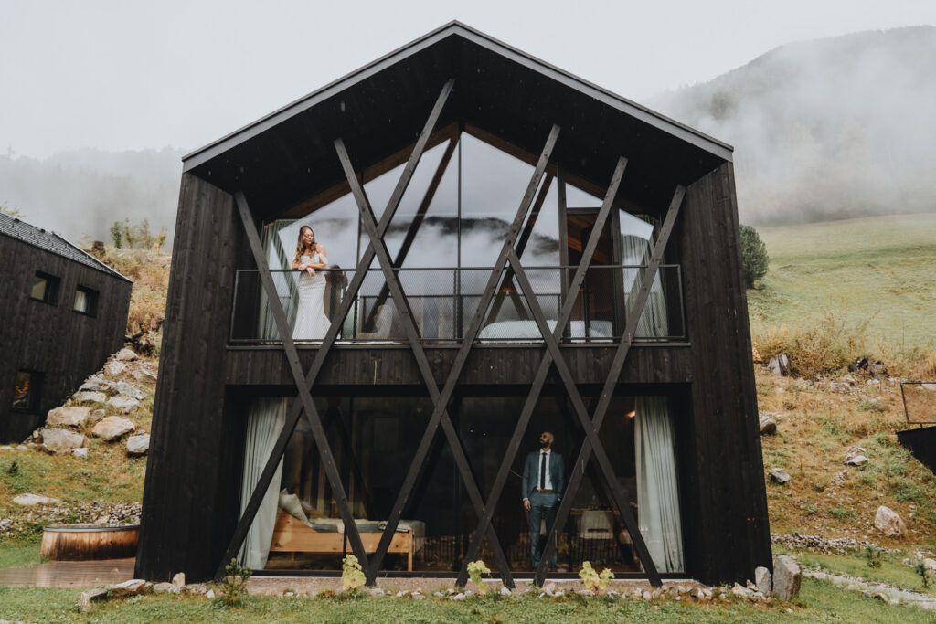 A couple in wedding clothes stand on separate floors of their Chalet in the Dolomites on their elopement day