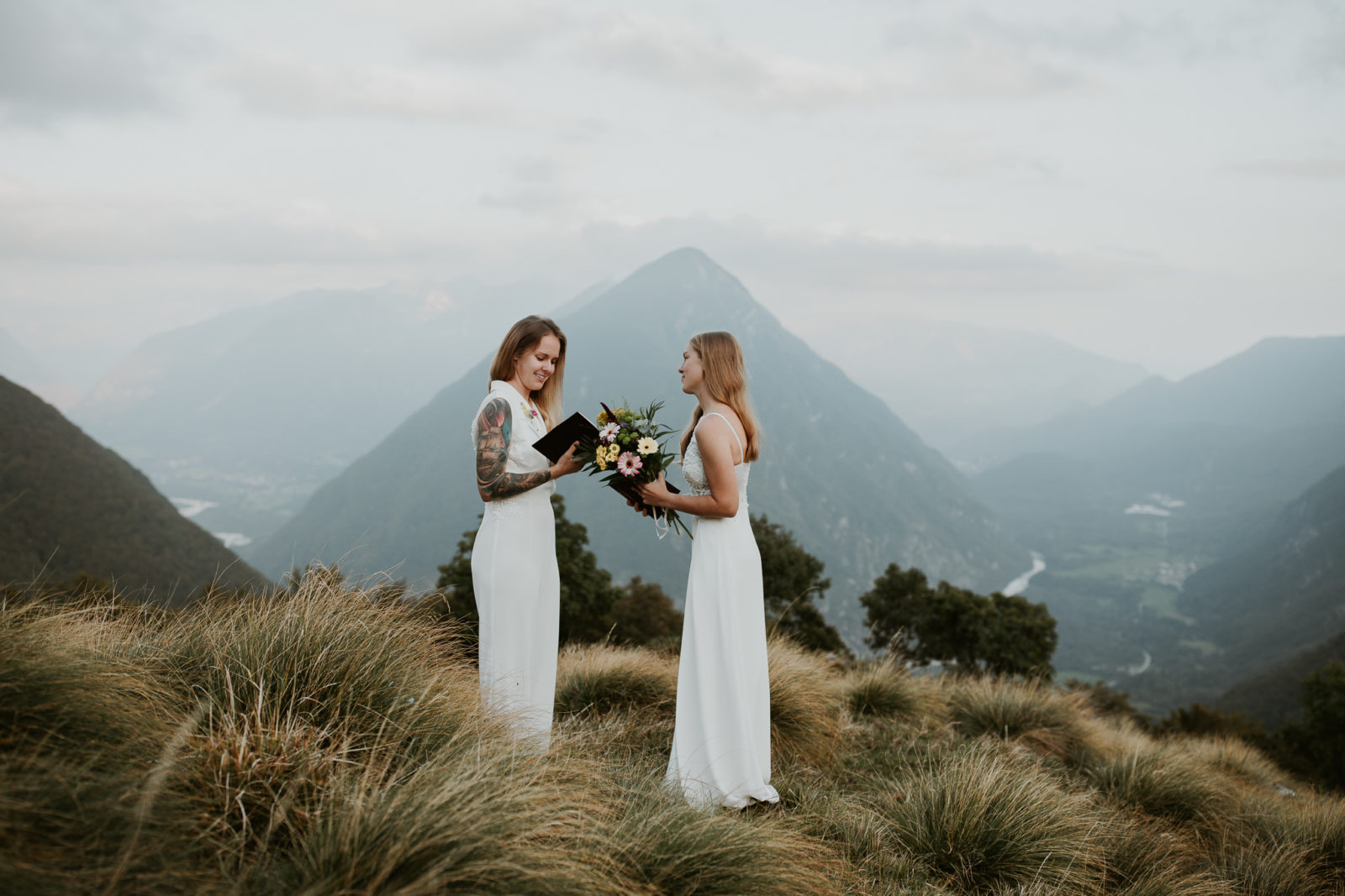 A lesbian couple stands reading their elopement vows in Triglav National park, with soft grasses in the foreground and tall mountains and the Soča River in the distance.