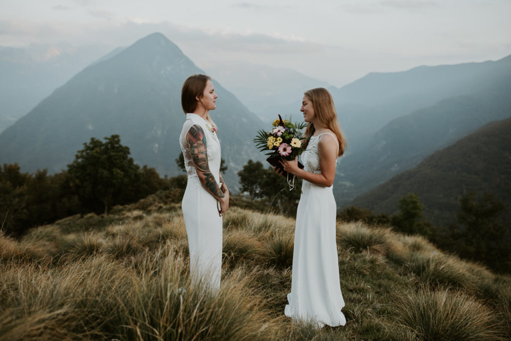 Lesbian couple holds flowers and vow books as they read their vows near sunset during their slovenia elopement photoshoot