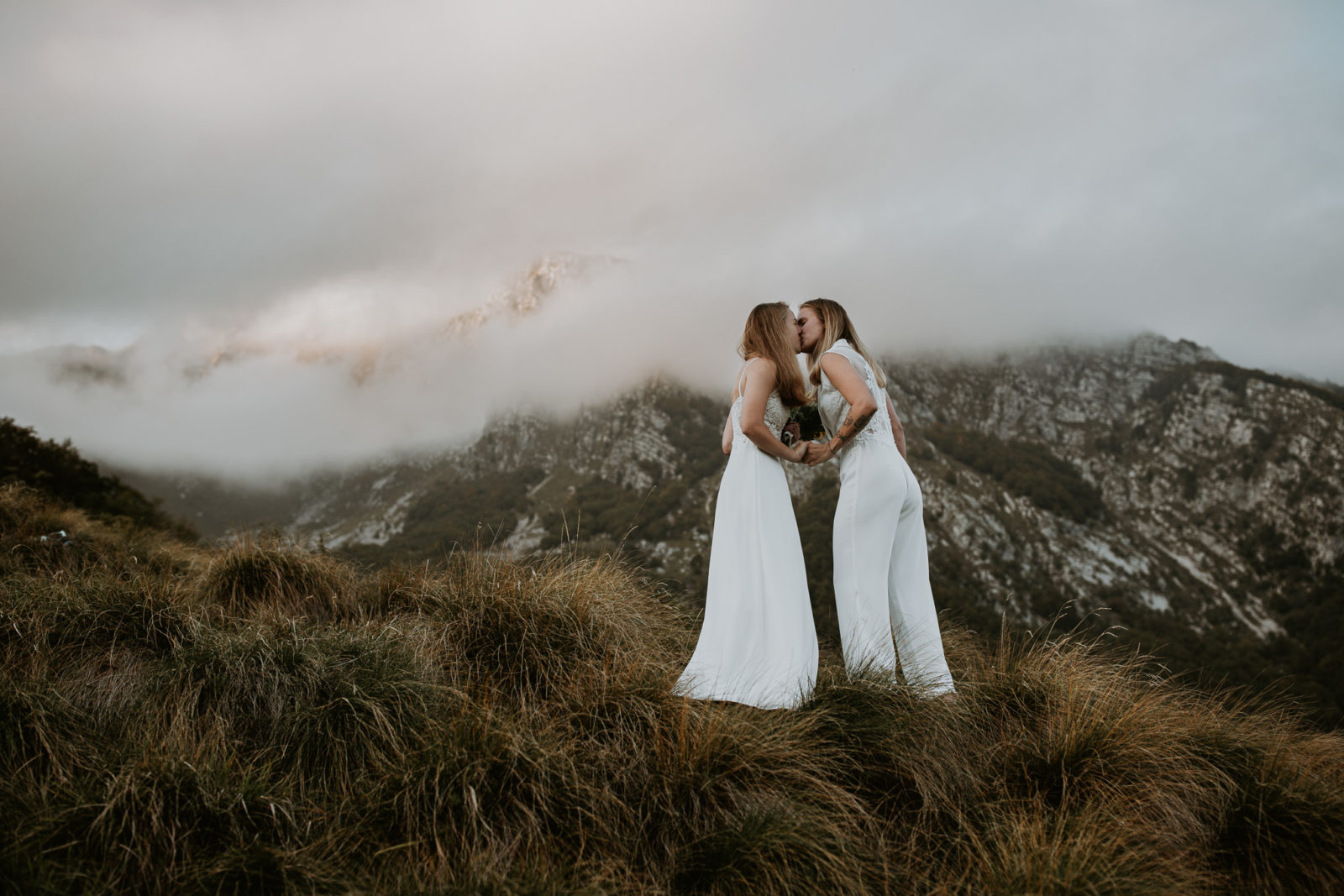 A lesbian couple in wedding dresses share a kiss with dramatic mountains in the background on their Slovenia elopement day
