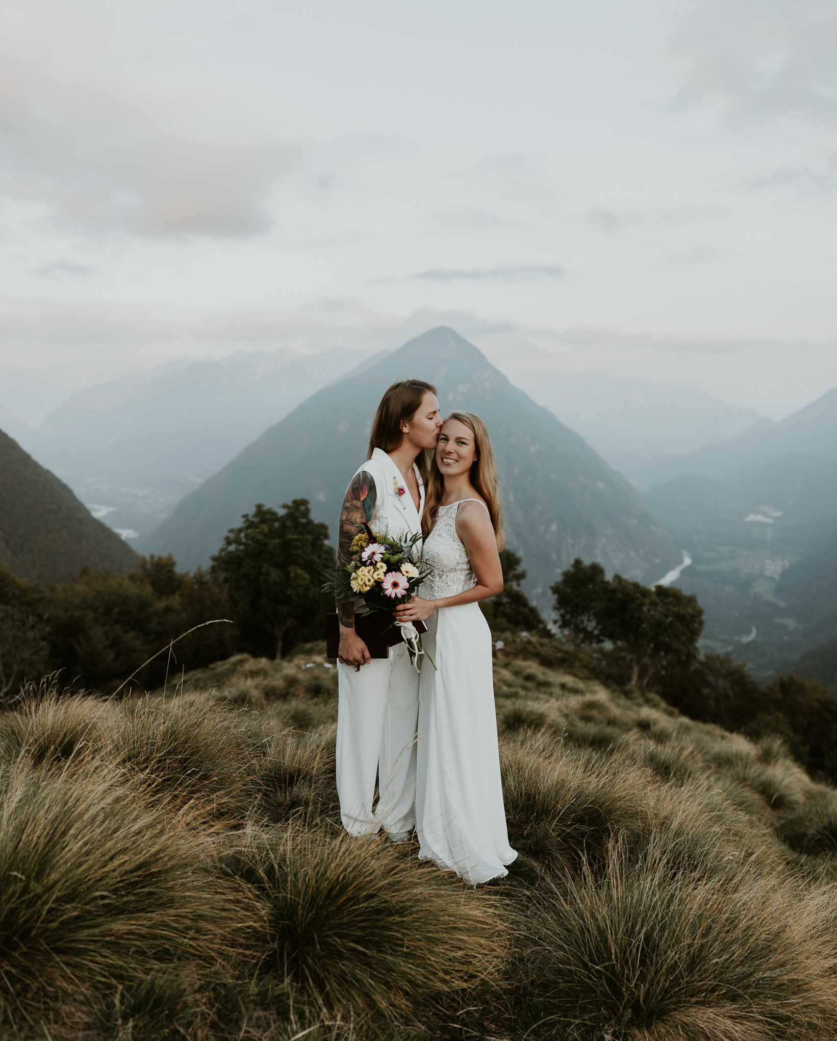 A couple kisses and smiles at the camera during their hiking elopement in the Slovenian Alps