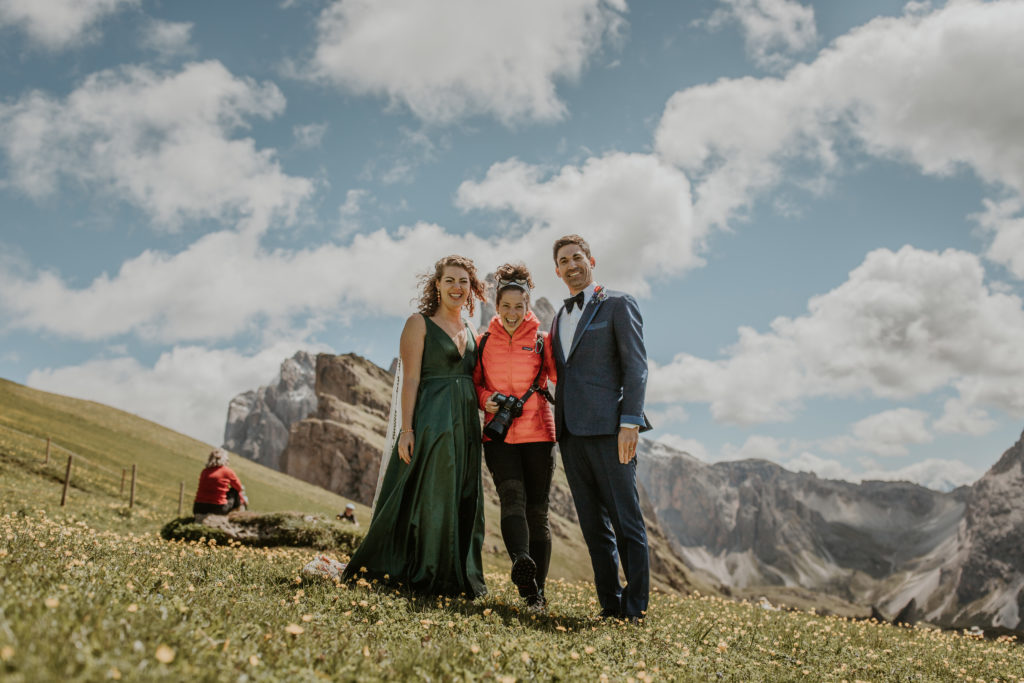 dolomites elopement photographer mariah arianna stands with two clients smiling and laughing into the camera at the top of Seceda, in the Italian Dolomites.