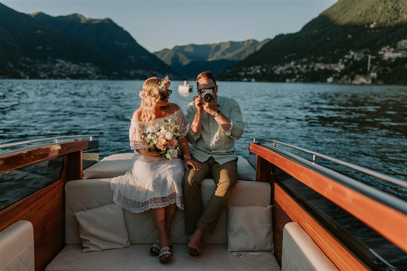 This is an image of a couple sitting on a boat on Lake Como during their elopement day. The man is holding a polaroid camera to his face, toward the camera, and his wife is smiling at him.