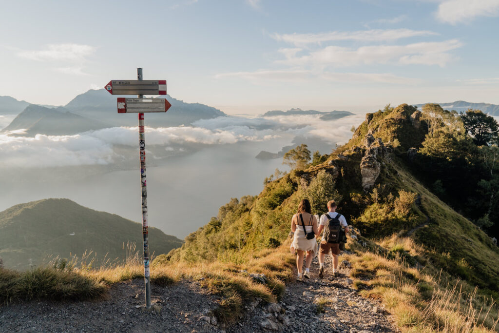 A wedding couple hike down a mountain trail with far off views of Lake Como in front of them on their elopement day