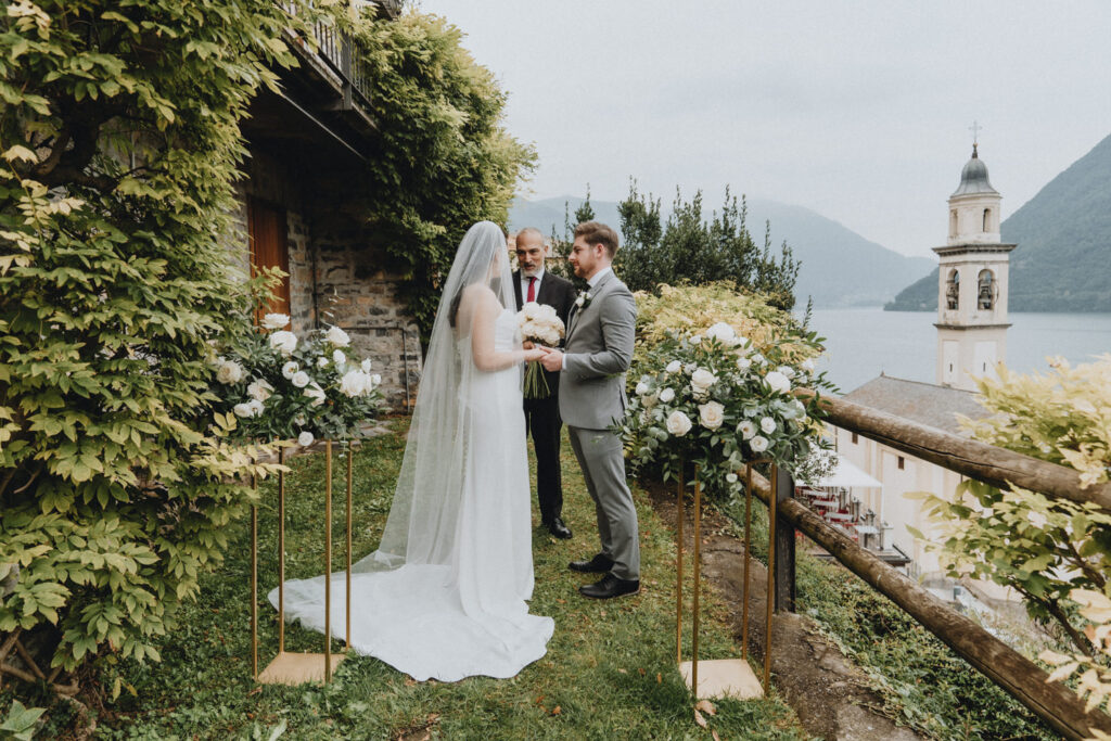 An eloping couple waring white dress and grey suit hold hands while a celebrant officiates their ceremony, surrounded by white flowers and Lake Como's classic lake and mountain views.