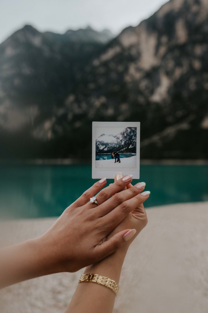 A womans' hands are seen holding a polaroid of her fiance proposing at Lago di Braies. Her ring is visible with the mountain lake in the background.
