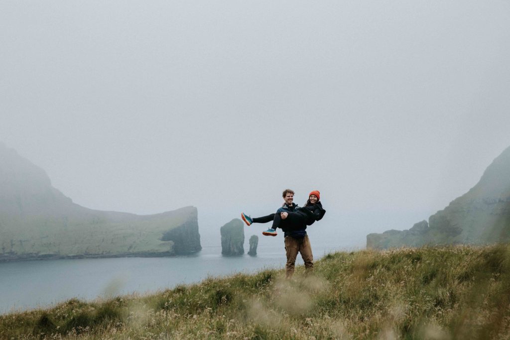 couple elopes in the Faroe Islands. They are in a field overlooking the ocean. The man is carrying the woman and they are both laughing.