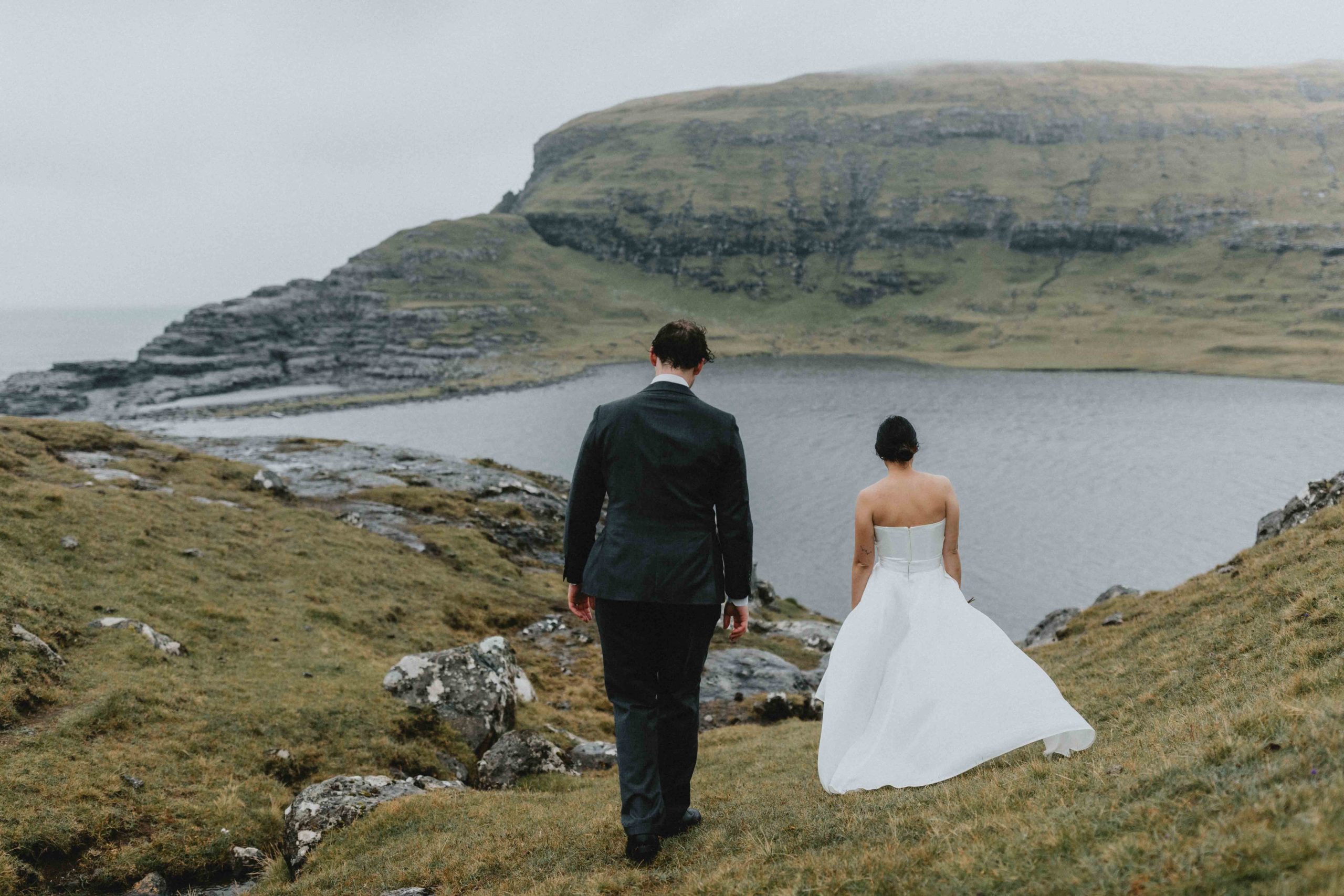 This image is of a couple walking away from the camera wearing wedding attire. They are walking toward Sorvagsvatn in the Faroe Islands. it is lightly raining and the wind is blowing.