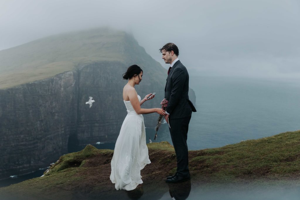 A wedding couple stands on a dramatic sea cliff reading their vows to one another. There is a bird flying in the background and it is misty and moody.