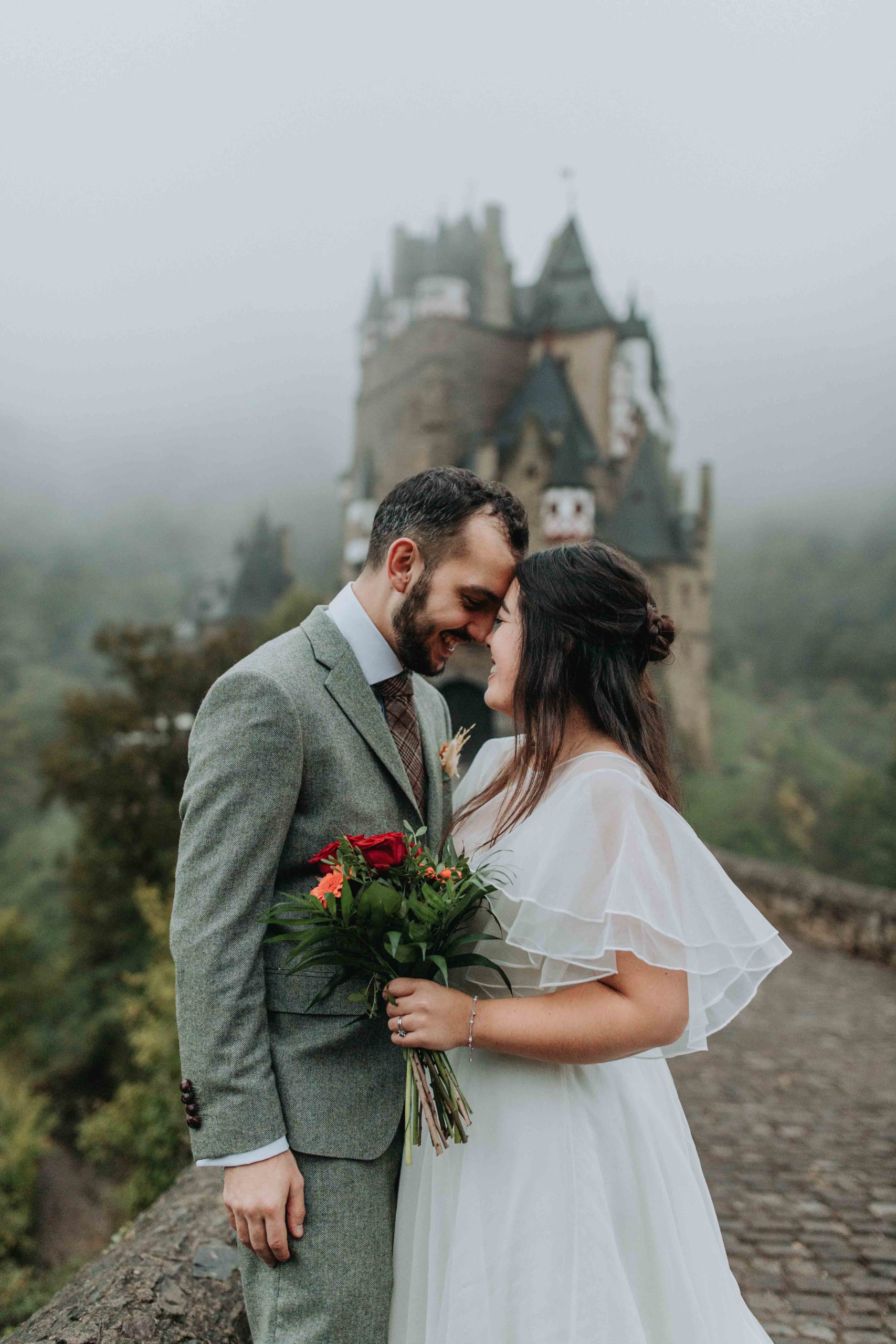Couple stands forehead to forehead smiling outside of Brug Eltz castle in Germany during their wedding