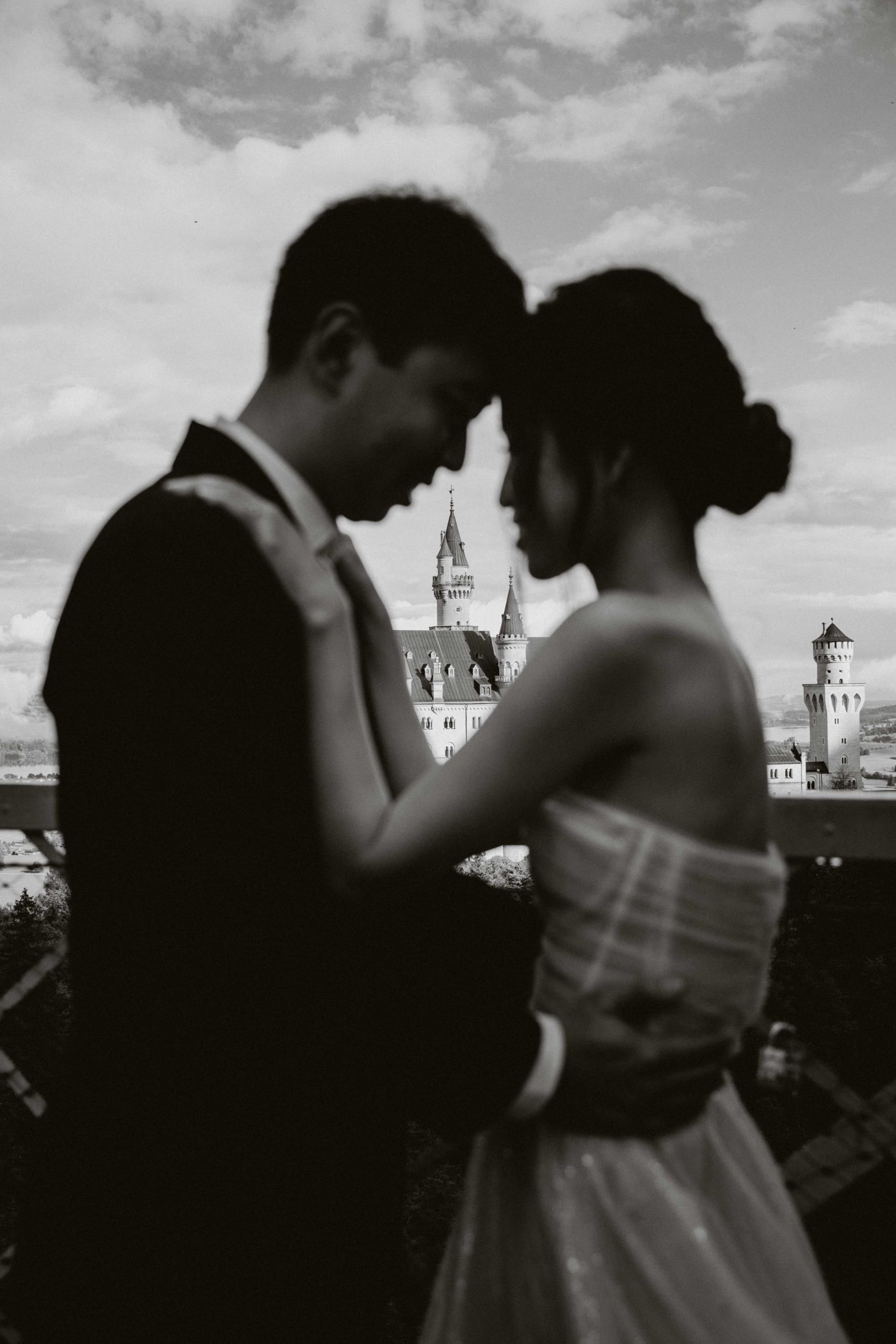 This is a black and white image of a wedding couple standing forehead to forehead with Schloss Neuschwanstein in the background between them