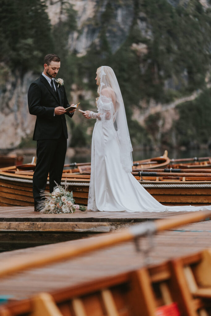 A couple wearing white wedding dress and dark suit read vows among wooden boats at a location in the Italian Dolomites 