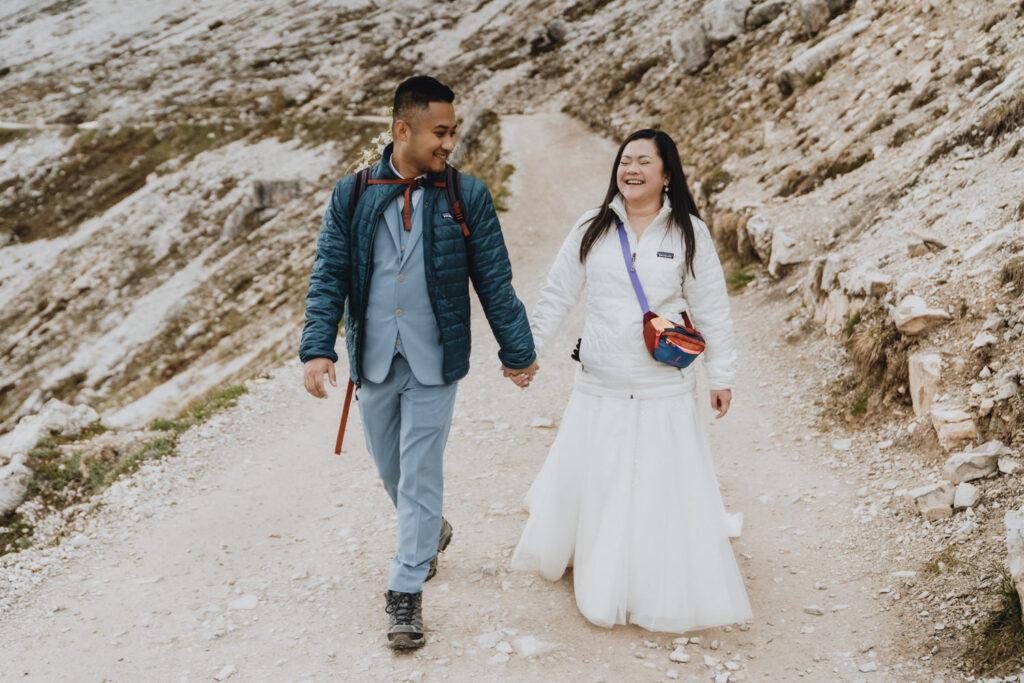 A couple is laughing during their elopement in the Dolomites, wearing a white Patagonia jacket and white wedding dress, and blue jacket and blue suit.