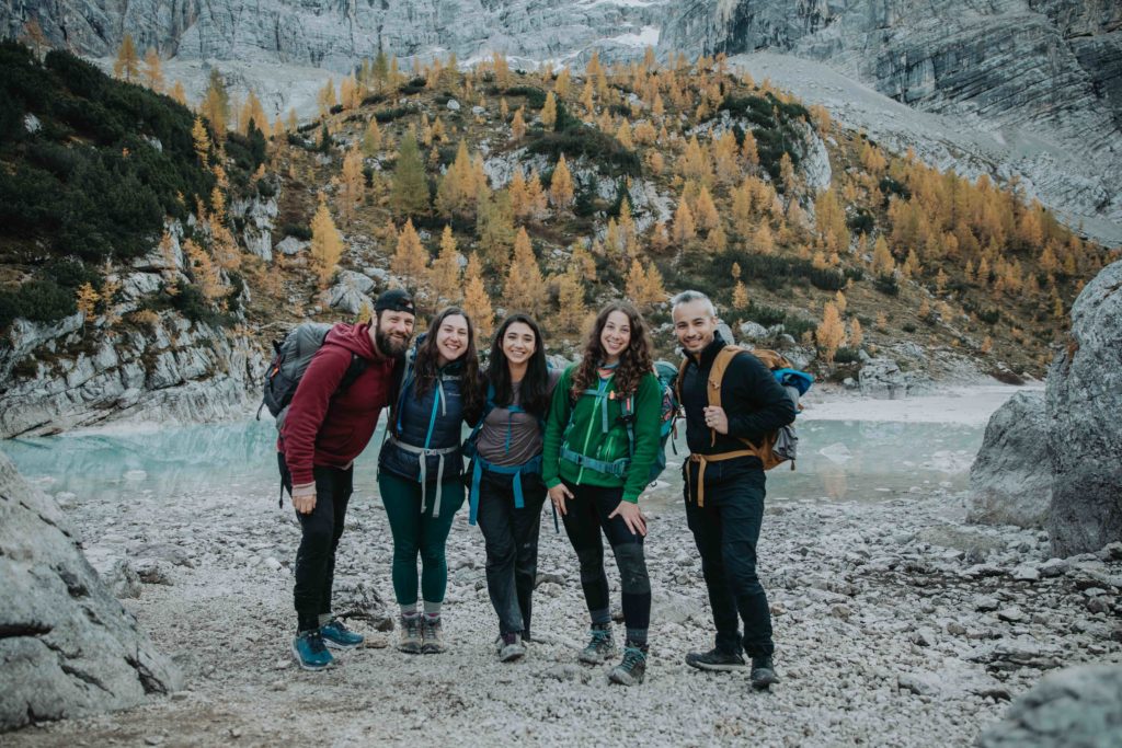 This is an image of a group of elopement guests standing next to Lake Sorapis during a couple's Dolomites hiking elopement. They are all standing in a line and smiling at the photo.