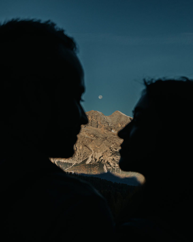 A stands facing each other with only their faces in silhouette, the moon and a mountain ridge in focus behind them.
