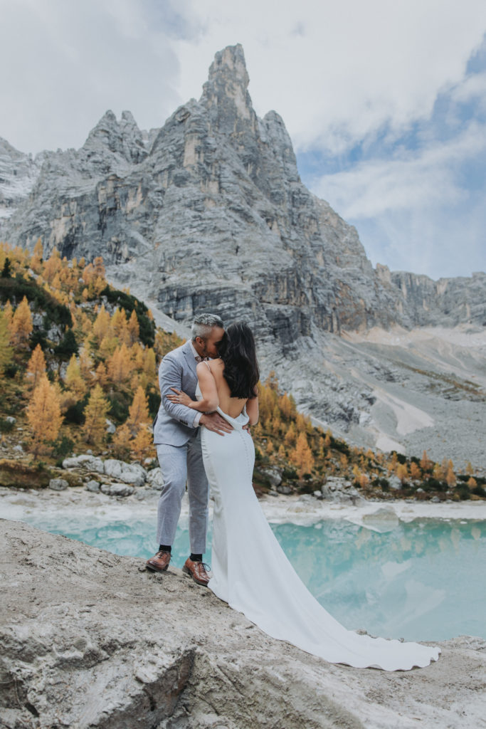 A couple is standing near Lake Sorapis during their 2 day Dolomites Italy elopement. The water is bright blue and the leaves golden. 