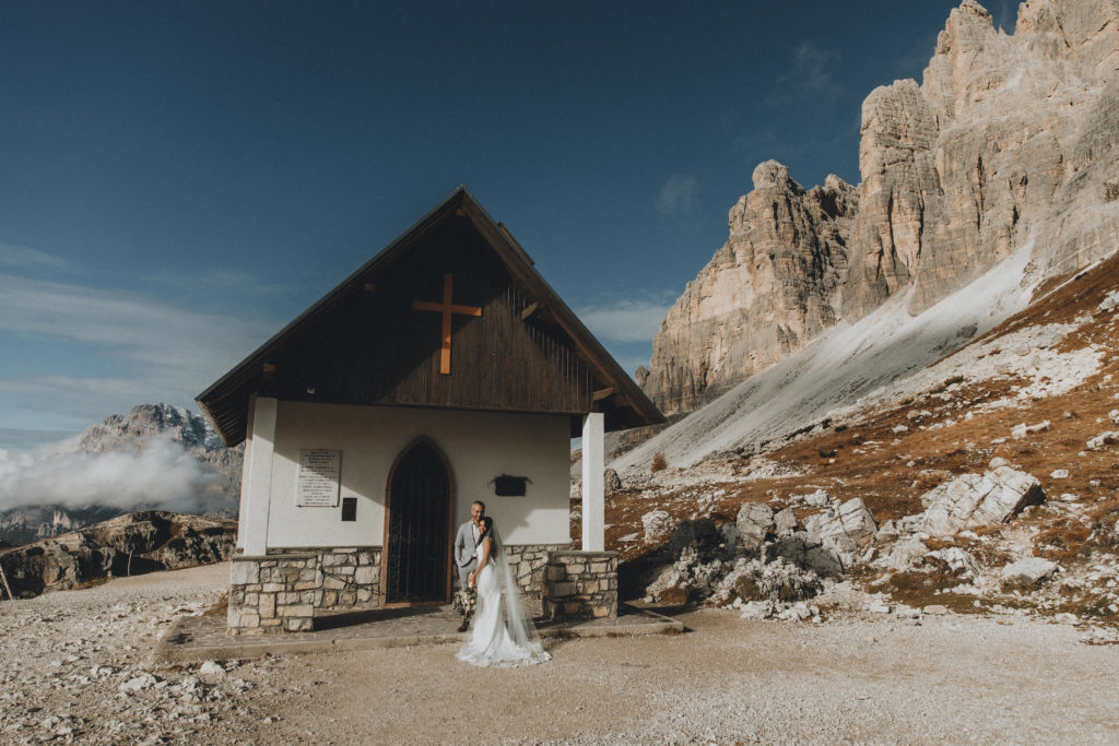 A couple wears their wedding attire next to a small mountain church during their Dolomites elopement.