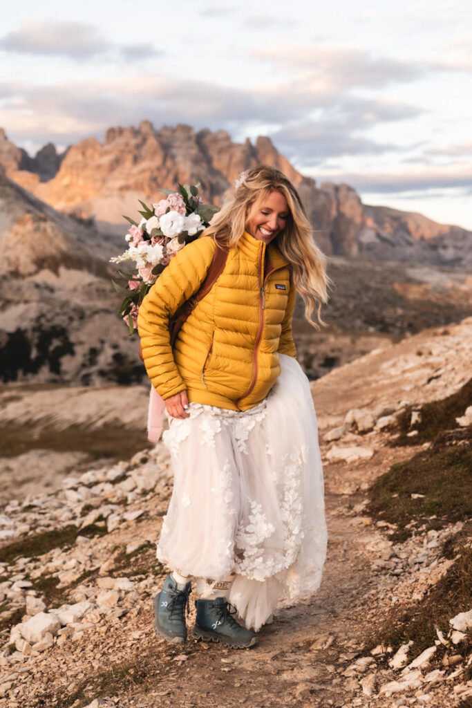 A bride wears a yellow Patagonia jacket over her wedding dress while hiking in the Dolomites on her elopement day