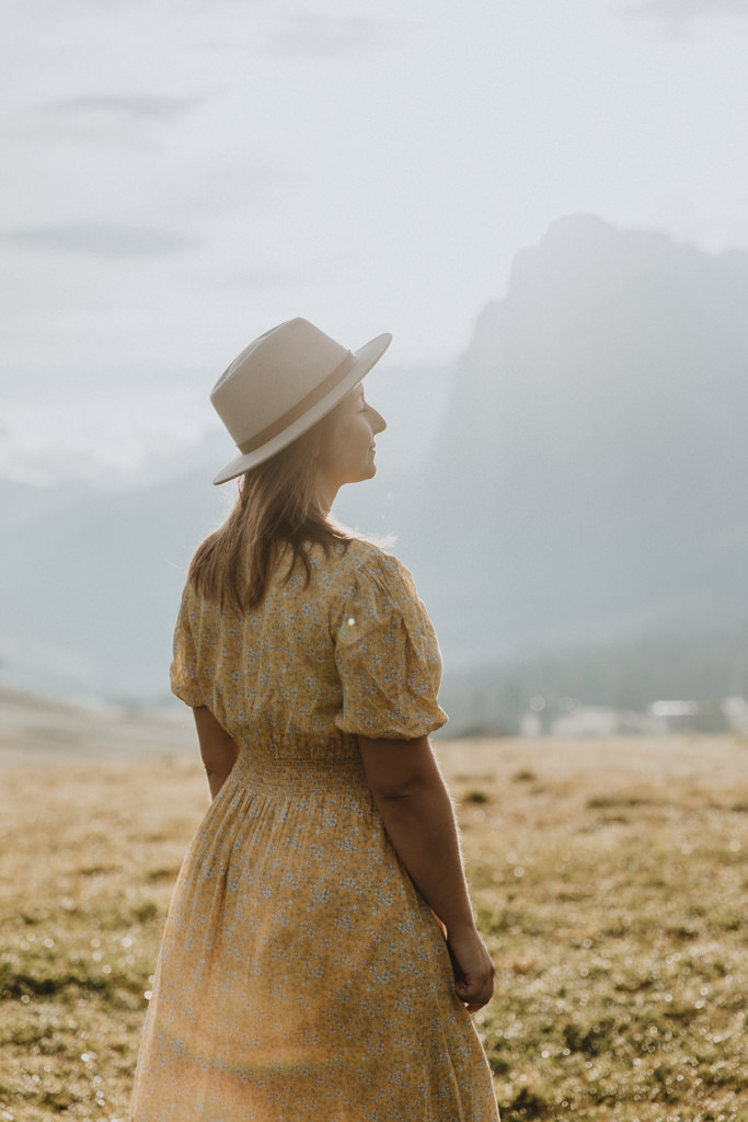 A woman stands in a yellow hat and sundress at Seiser Alm in the Dolomites. Her eyes are closed and the sun is bright and flaring against the lens.