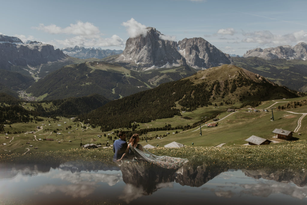 Couple wears their wedding attire during Dolomites elopement. They are sitting in a grassy green field with yellow flowers looking away at the mountains and huts across the valley.