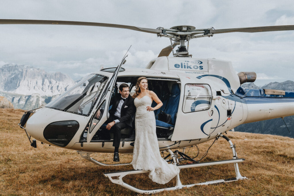 A couple stands in the door of a helicopter wearing a wedding dress and black tuxedo on their elopement day in the Dolomites