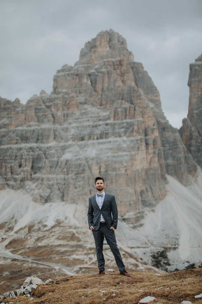 A man standing in a dark blue suit standing on a hill with a towering mountain peak behind him.