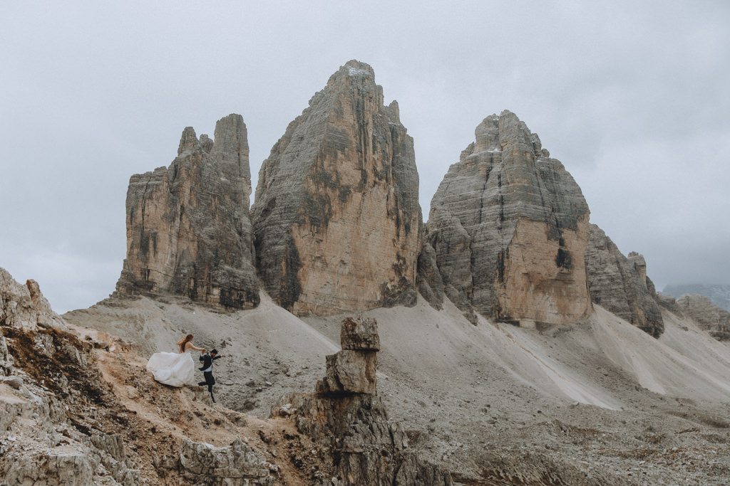 A couple wearing their wedding attire for their Dolomites adventure elopement runs down a rocky trail with the Tre Cime peaks framed behind them.