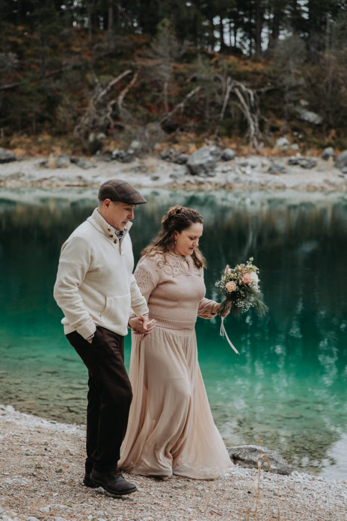 A couple in a dusty rose colored dress and beige sweater walk along Eibsee during their winter elopement in Germany