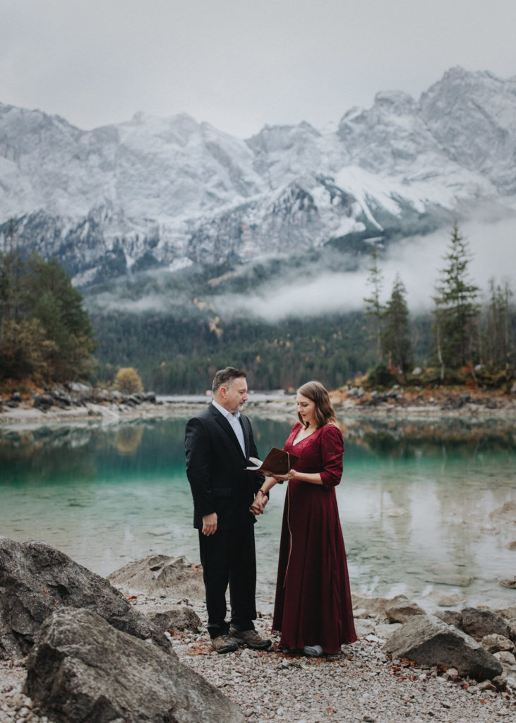 A couple stands facing each other in wedding attire holding a vow book as they elope in Germany, near Eibsee.