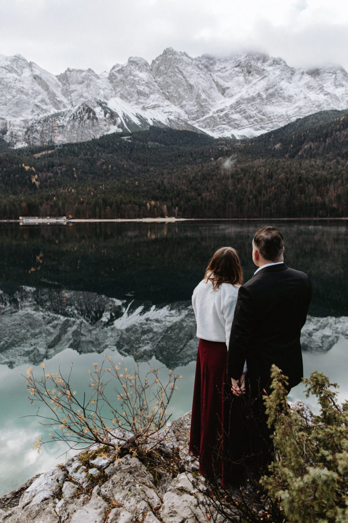 A wedding couple stands near Eibsee as they elope in Germany. They are standing with their backs to their camera looking at a snow covered mountain reflected on the lake.
