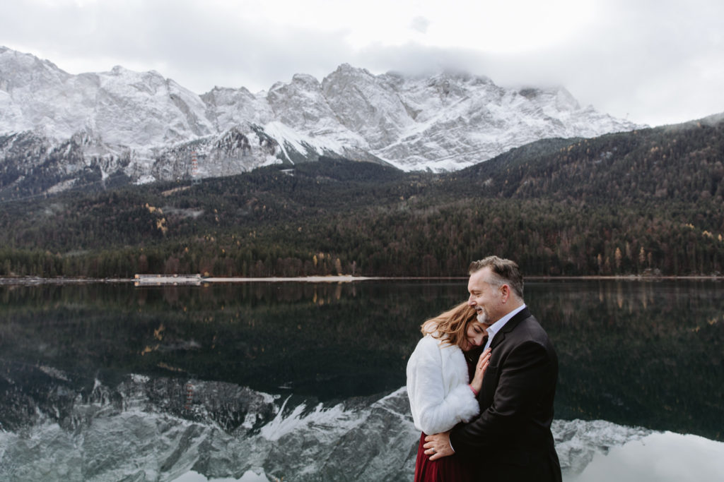 middle aged couple eloping in Austria and Germany, Eibsee. Couple is standing chest to chest and there is a lake reflecting snow covered mountains behind them.