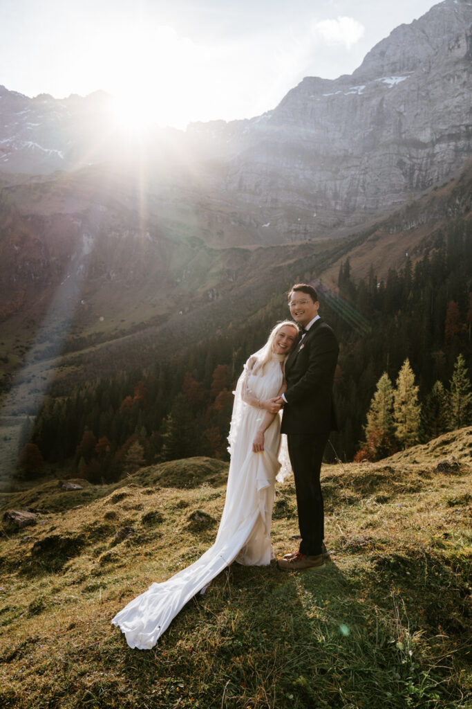A couple stand in white wedding dress and black tux, backlit by the sun coming over high grey mountains at this outdoor elopement location in Germany