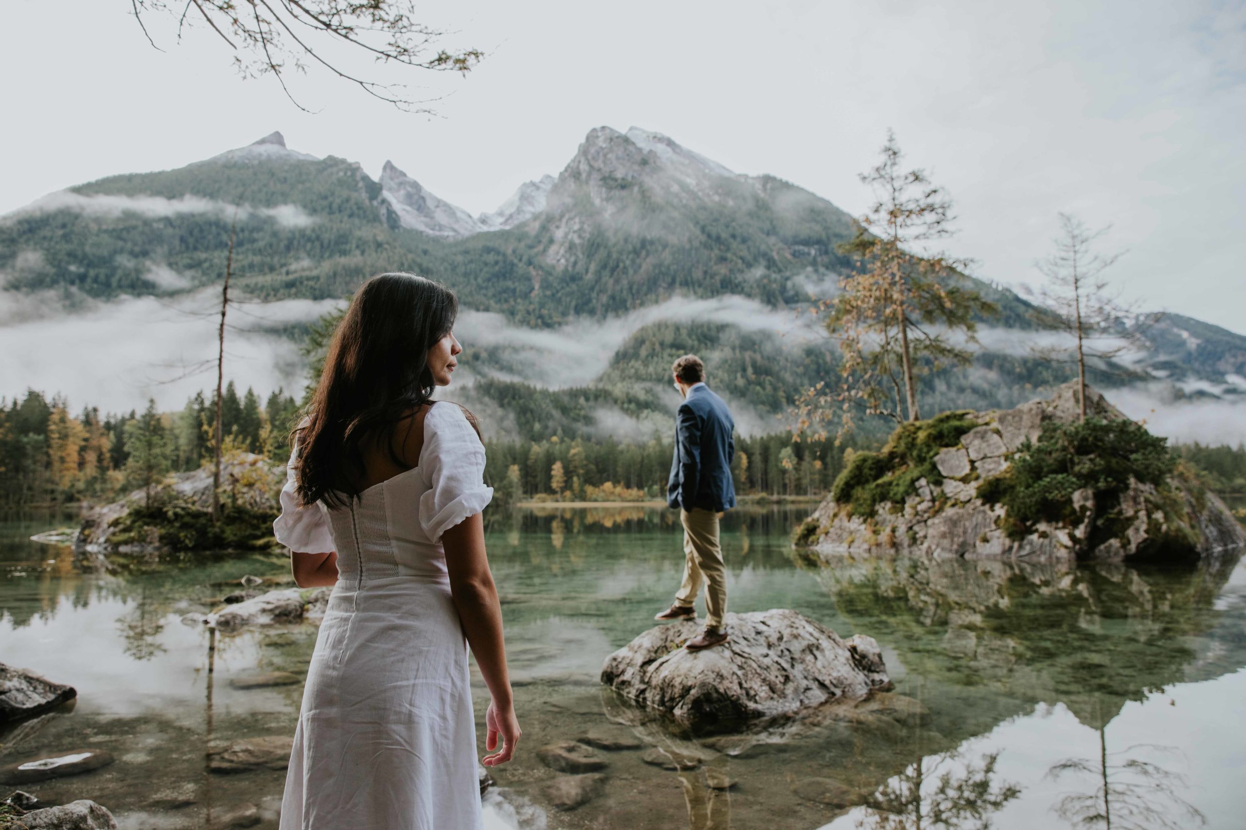Berchtesgaden, Germany elopement photos of a couple standing on large stones in the water with mountains in the background. She is facing to the side and he has his back to the camera.