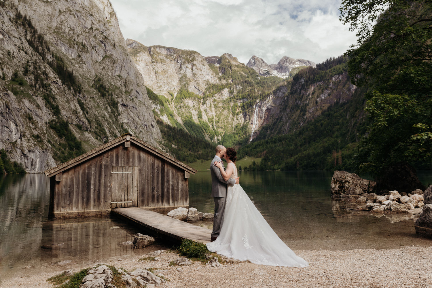 A couple embrace in a wedding dress and grey suit next to a still lake and wooden fishing cabin in the German Alps, a popular elopement location in Germany