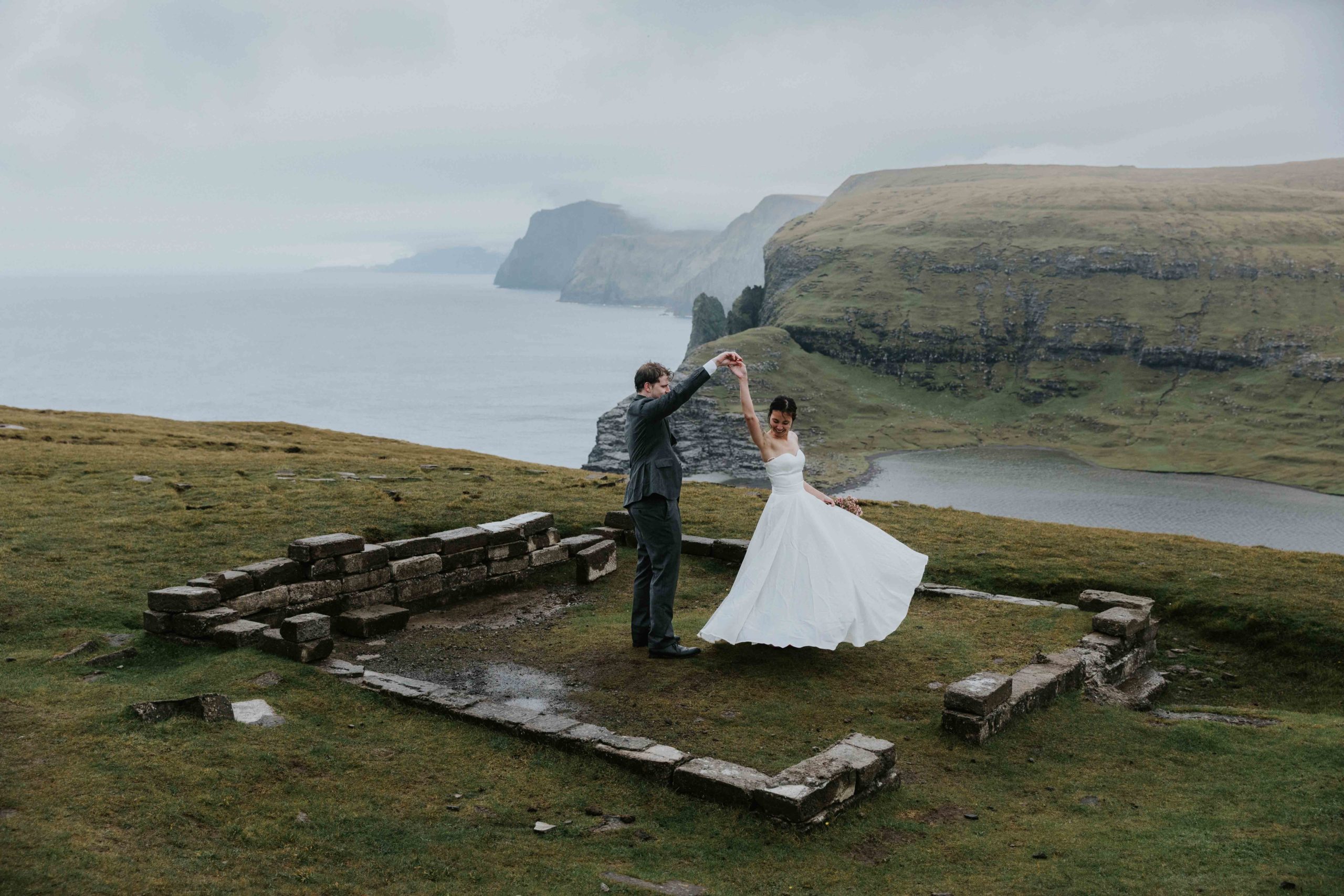 Moody photo of a Faroe islands elopement. There is a couple wearing wedding attire dancing and spinning in old stone ruins with sea cliffs in the background.
