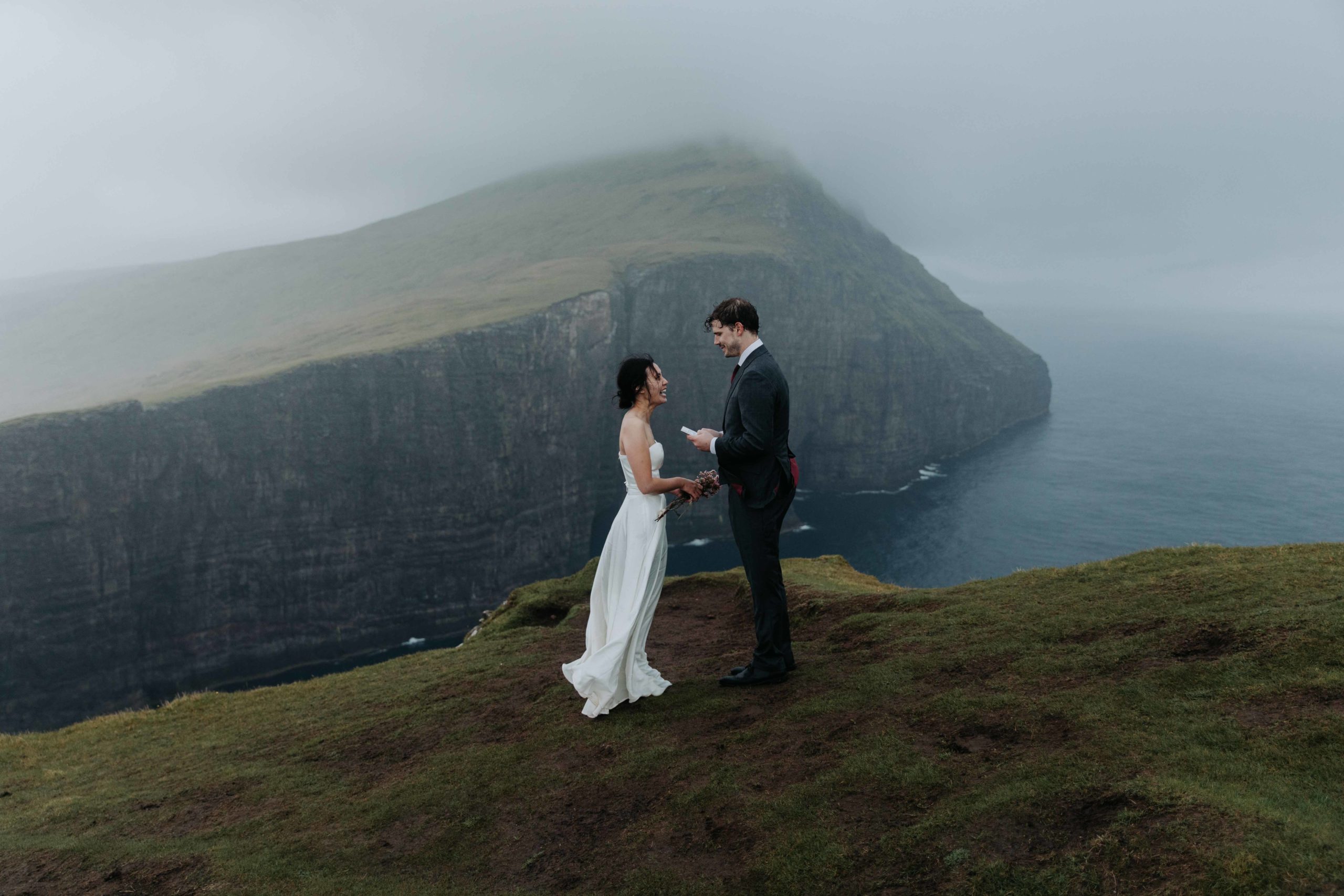 A couple is standing next to dramatic sea cliff on the Faroe Islands reading their wedding vows. They are facing each other and smiling.