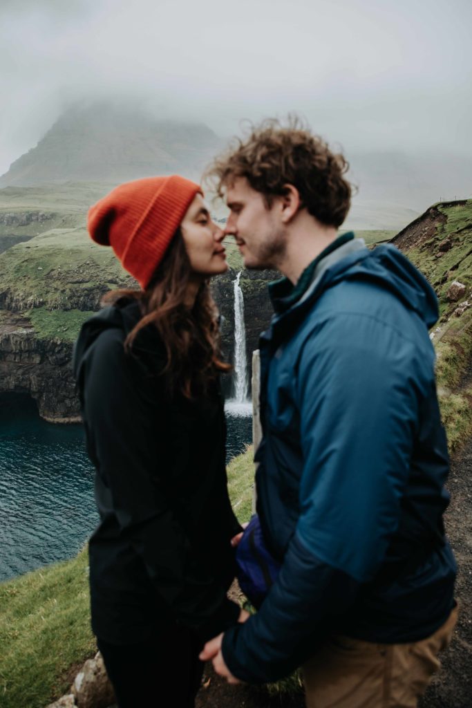 A couple stands pressing their noses together with their eyes closed near the Mulafossur on the Faroe Islands. 