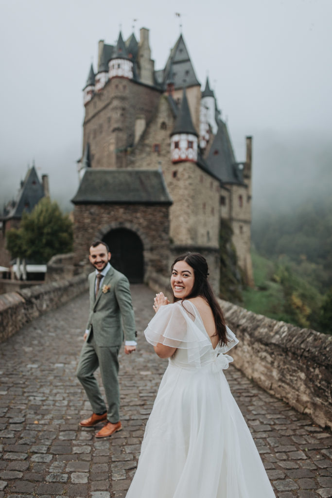 A couple looks at the camera laughing in their wedding clothes outside Burg Eltz in Germany