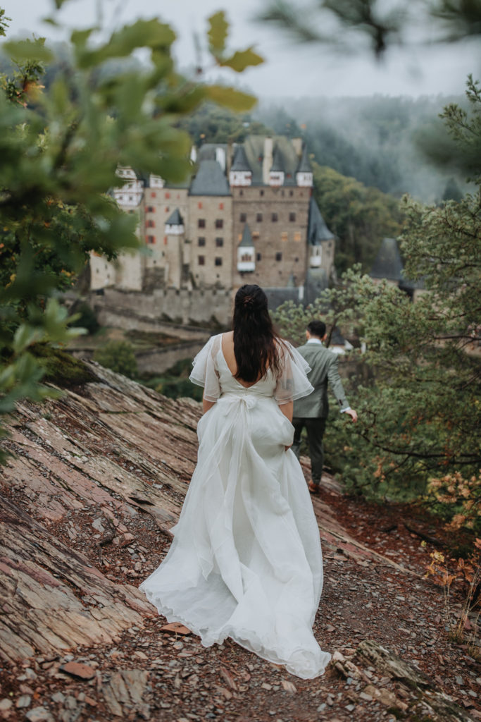 A woman in a white wedding dress walks away from the camera toward Burg Eltz in Germany