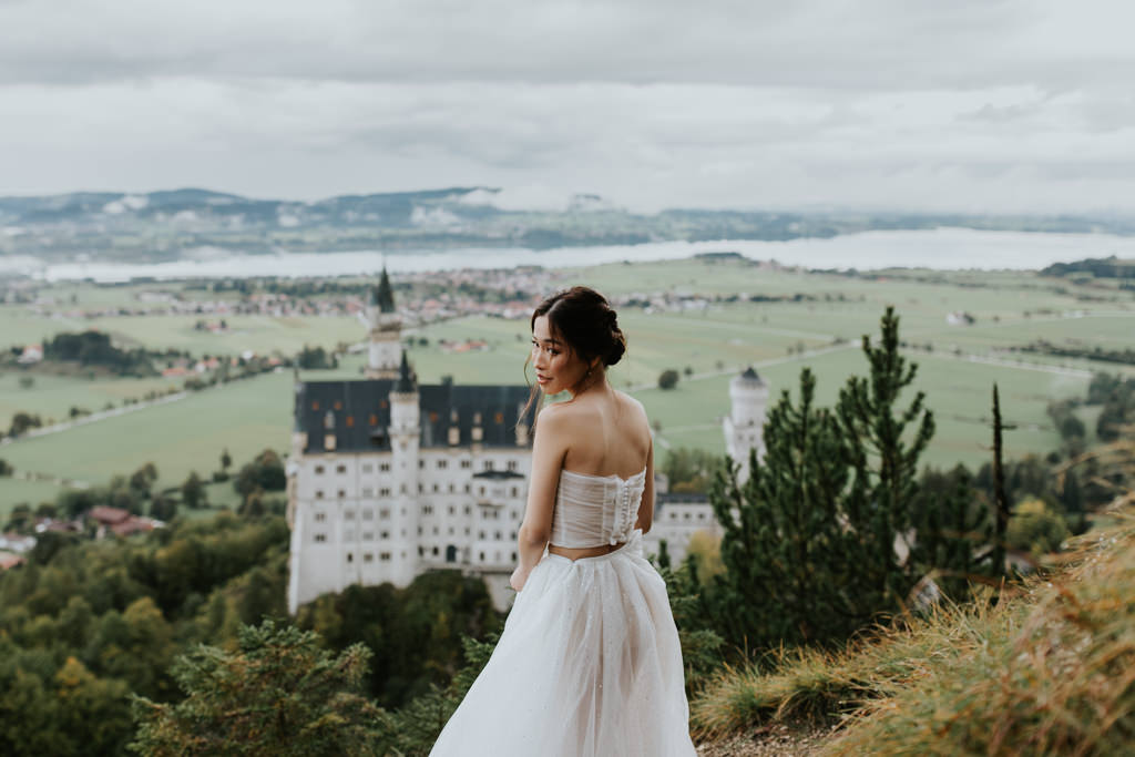 A petite asian woman looks over her shoulder wearing a white wedding gown with the impressive Schloss Neuschwanstein in the background.