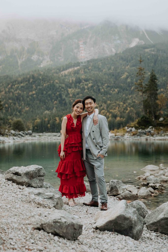 An asian couple stands smiling at the camera wearing a red dress and grey suit next to a lake in the alps for a couples photo session