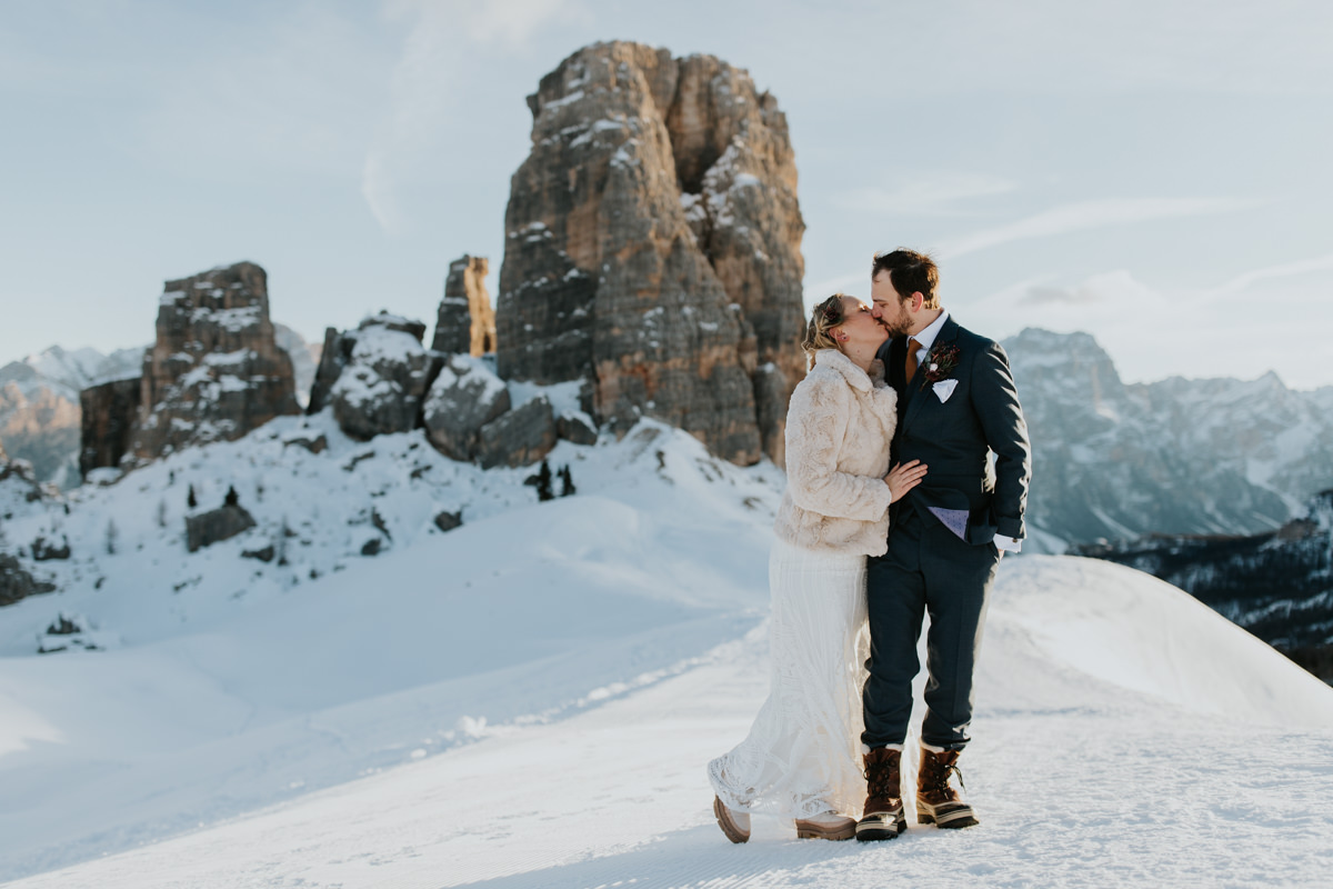 A couple in winter wedding attire stand on a mountain top in the Italian Dolomites for their elopement. They are standing facing each other kissing. The sun is shining and the mountains are covered in snow.