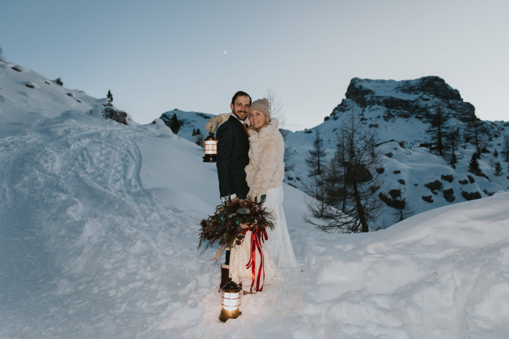 Wedding couple during their Dolomites ski wedding near Cinque Torri stand embracing, smiling at the camera. It is after sunset and the moon is in the sky above them. They are holding glowing lanterns and big bouquet.