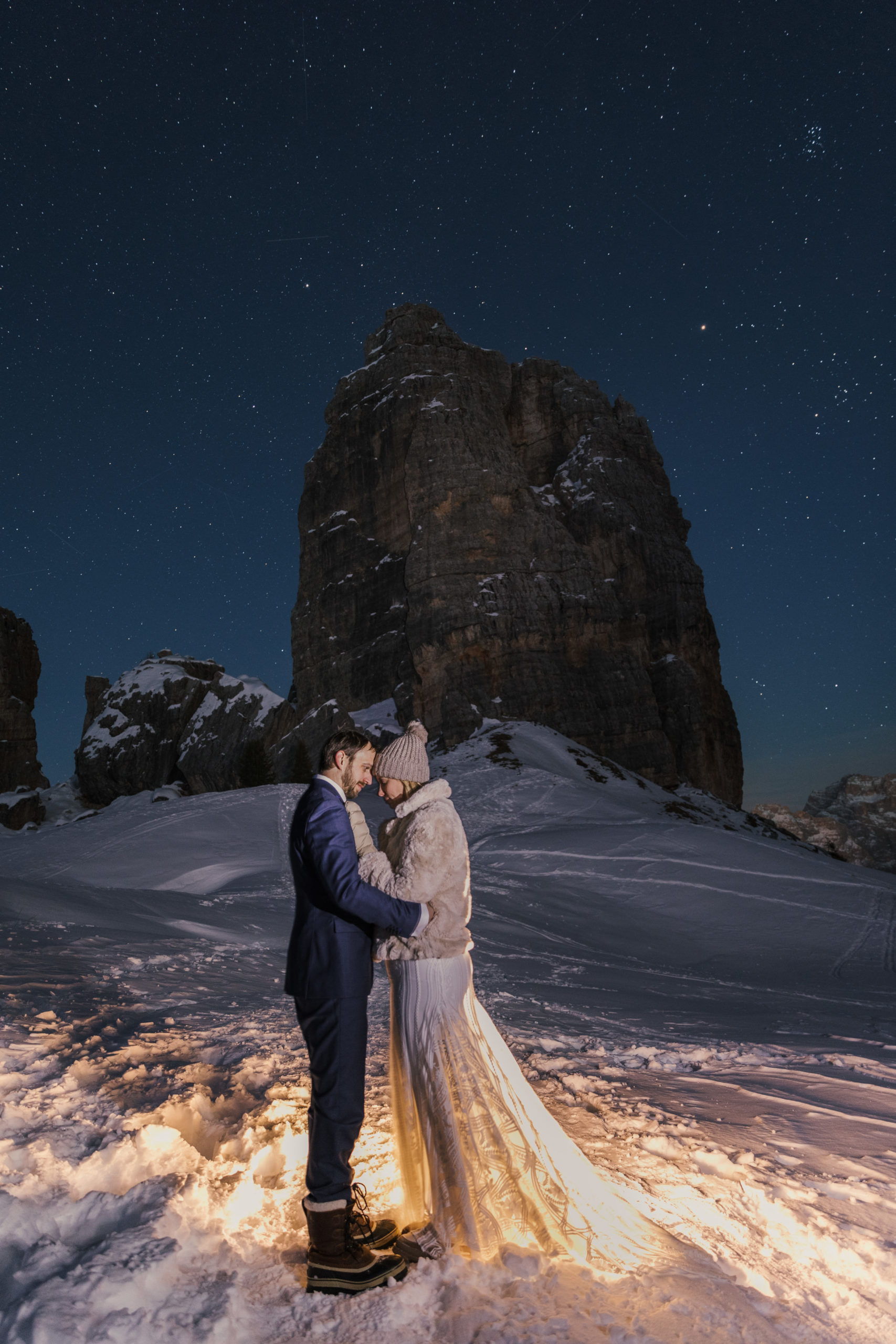A couple pose for winter elopement photos in the Dolomites, with a starry night skh above them and jagged mountains behind them.