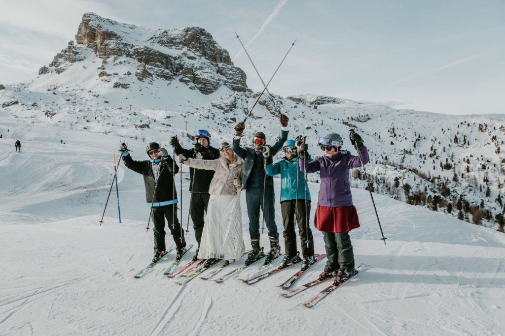 A family stands in a line on the slopes wearing skis and wedding attire for a Dolomites ski wedding. They are holding their ski poles up and cheering.